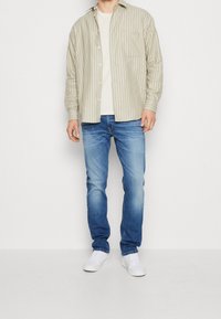 Camisa a rayas beige claro, camiseta crema, vaqueros azul y zapatillas blancas. La camisa tiene botones y un bolsillo en el pecho; los vaqueros están desgastados.