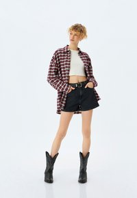 Punainen ja valkoinen ruudullinen oversized-paita, valkoinen lyhyt toppi, mustat farkkushortsit, mustat cowboy-kengät tekstuurilla, seisomassa vaaleaa taustaa vasten.