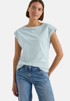 Femme aux cheveux bruns courts, portant un haut sans manches bleu clair et un jean bleu, debout avec une main sur la hanche sur un fond clair.
