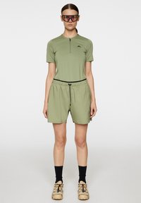 Oliivinvihreä lyhythihainen paita, jossa on puolivetoketju, ja siihen sopivat shortsit, jotka sisältävät joustavan vyötärönauhan ja nauhakuulokkeita, yhdistettynä vaaleanbeigeihin kenkiin.