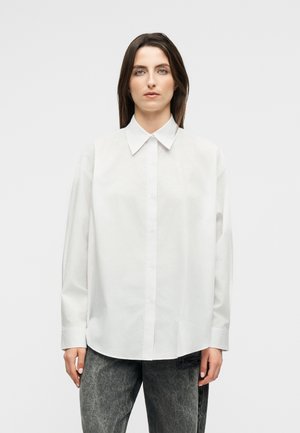 Žena v bielej voľnej košeli s gombíkmi a špicatým golierom, oblečená v čiernych vypraných džínsoch, stojaca pred jednoduchým svetlým pozadím.