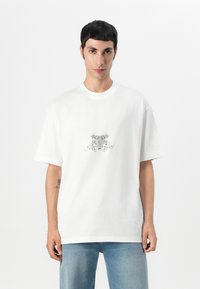 T-shirt blanc en coton avec une coupe décontractée, doté d'un motif graphique gris au centre. Manches courtes et col rond.