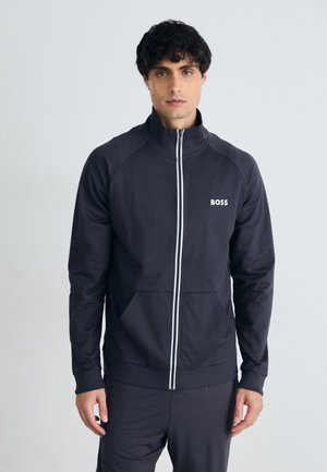 Schwarze Zip-Jacke mit hohem Kragen, versehen mit weißem Logo und Streifen. Das Material wirkt glatt; das Design umfasst zwei Fronttaschen.