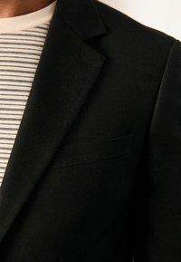 Blazer negro con solapa de muesca, tejido texturizado y un bolsillo en el pecho, superpuesto a una camisa ligera a rayas.