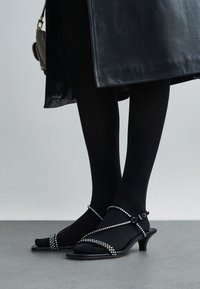 Czarne sandały na obcasie o eleganckim designie, z błyszczącymi zdobieniami na paskach i szpiczastym noskiem, noszone z czarnymi rajstopami.