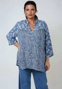 Woman with dark hair wearing a blue long-sleeve blouse with floral and paisley patterns and blue pants, standing against a plain background.