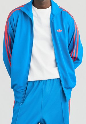 Persoon die een felblauw trainingspak met roze strepen en een wit T-shirt draagt, staat met de handen achter de rug.