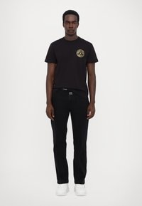 Tricou din bumbac negru cu guler rotund și detaliu cu logo auriu pe partea stângă a pieptului, asortat cu blugi negri și adidași albi.