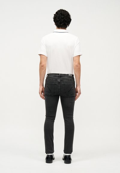 Göndör hajú férfi háttal áll, fehér rövid ujjú pólót, fekete szűk farmernadrágot, fehér zoknit és fekete elegáns cipőt visel.