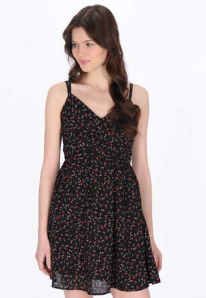 Kobieta ubrane w czarną sukienkę bez rękawów z drobnym czerwonym wzorem kwiatowym, o luźnym kroju i marszczonym pasie, patrząca na bok z długimi falowanymi włosami.