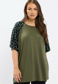 Vrouw in een olijfgroene losse top met zwart en olijfgroen luipaardprint op korte mouwen, staand tegen een effen witte achtergrond.