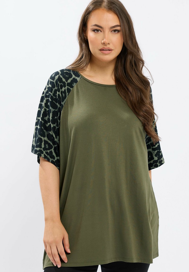 Vrouw in een olijfgroene losse top met zwart en olijfgroen luipaardprint op korte mouwen, staand tegen een effen witte achtergrond.