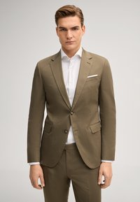 Mann in olivgrünem Anzug und weißem Hemd, der vor einem einfarbigen hellen Hintergrund nach vorn blickt.