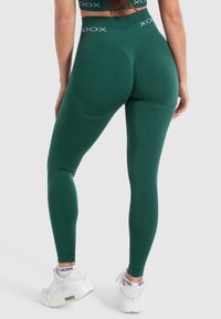 Groene leggings gemaakt van rekbaar materiaal met een hoge tailleband met een logo. Het ontwerp is figuuraccentuerend en naadloos aan de achterkant.