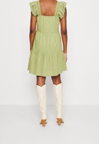 Lichtgroene mouwloze jurk met kanten rouches aan de bandjes, een gelaagde rok en een vierkante rug. Gecombineerd met kniehoge witte laarzen met hak.