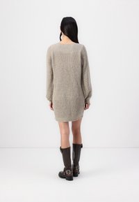 Femme aux longs cheveux foncés portant une robe-pull tricotée beige clair et des bottes hautes marron foncé, debout de dos sur un fond blanc uni.
