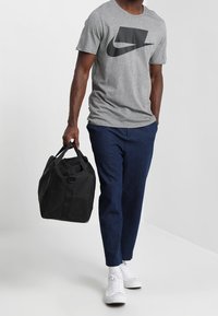 Šedé tričko s krátkým rukávem s černým logem Nike, modré zúžené kalhoty, bílé tenisky a černá sportovní taška s uchy.