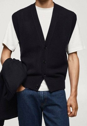 Mand iført sort knaplukket strikvest over hvid kortærmet skjorte, blå jeans, holder en sort jakke i venstre hånd.