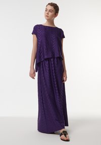 Paarse two-piece jurk met een gestructureerde top met een gelaagd ontwerp en een lange rok met een bladmotief. Gecombineerd met zwarte sandalen.