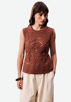 Femme portant un haut sans manches en tricot couleur rouille avec un motif géométrique en dentelle, un pantalon large crème, et un bracelet foncé orné de perles, regardant vers le bas.