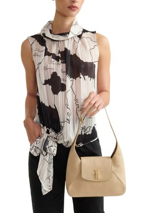 Donna con camicetta senza maniche con stampa di mappe annodata in vita, pantaloni neri, che tiene una borsa beige con chiusura dorata sulla spalla.