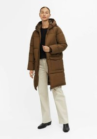 Doudoune longue marron avec capuche, dotée d'une fermeture éclair à l'avant et de poches plaquées. Associée à un pantalon beige clair et des chaussures noires.