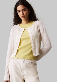 Weißer Cardigan mit langen Ärmeln, Knopfleiste; drüber getragen über einem gelben Rippenoberteil. Model trägt hellfarbene Jeans. Glatte Textur, lässiger Stil.