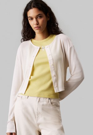 Weißer Cardigan mit langen Ärmeln, Knopfleiste; drüber getragen über einem gelben Rippenoberteil. Model trägt hellfarbene Jeans. Glatte Textur, lässiger Stil.