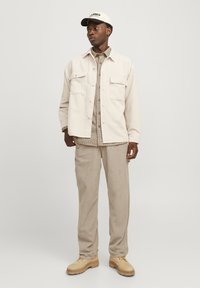 Beige corduroy jacket with two front pockets over a plaid shirt; paired with beige corduroy pants and light yellow boots. White cap.