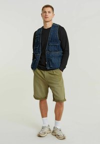 Gilet di jeans con più tasche, indossato sopra una maglietta nera a maniche lunghe, abbinato a pantaloni corti verde oliva arrotolati e sneakers crema.