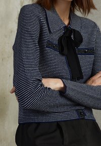 Laivastonsininen ja valkoinen houndstooth-kuvioinen takki mustilla yksityiskohdilla, jossa on solmuke ja napit, teksturoitu kangas, lyhyt malli.