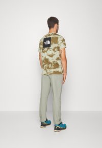 Muž stojící zády, oblečený v tričku s camo vzorem od North Face v zelené a béžové barvě, v světle šedých kalhotách a modrých turistických botách.