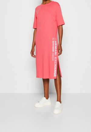 Person iført en koral midi kjole med korte ærmer og hvide sneakers, med lodret hvid tekst på siden, der lyder Armani Exchange.