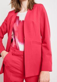 Mujer vistiendo un blazer rosa brillante y pantalones a juego con una blusa de patrón abstracto en blanco y rosa, con una mano en el bolsillo, contra un fondo liso.