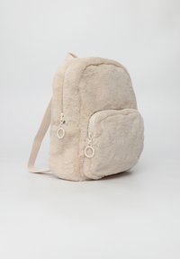 Beżowy plecak z futra syntetycznego o zaokrąglonym kształcie, z dwoma zamykanymi na zamek komorami i materiałowymi paskami. Miękka tekstura z delikatnym połyskiem.