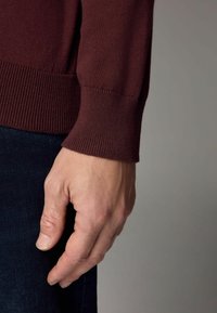 Burgundi pulóver bordás mandzsettákkal, sötét farmer anyag mellett megjelenítve, amelyen egy kéz pihen. Sima textúra, testhezálló dizájn.