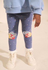 Collants bleus à pois roses, ornés d'un motif coloré de coccinelle sur les genoux, assortis à des chaussettes blanches à bordure en dentelle et des baskets blanches.