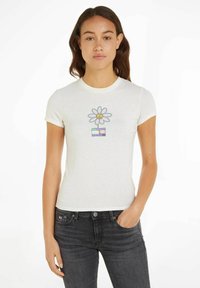 Camiseta blanca ajustada de manga corta, con un gráfico colorido de una flor y el texto "PAZ" debajo. Combinada con jeans gris oscuro.