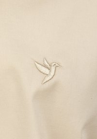 Tela beige con una textura suave que presenta un delicado diseño de colibrí bordado en beige. No hay otros patrones ni acentos presentes.