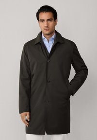 Tumma vihreä, pitkä takki, joka on valmistettu sileästä kankaasta, jossa on kauluskraagi, napitus ja sivutaskut, puettuna raitapaidan päälle.