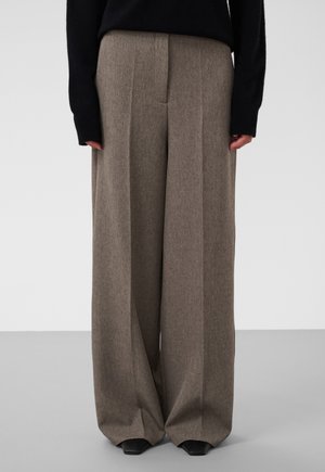 Weit geschnittene braune Hose mit vorderen Bügelfalten, getragen mit einem schwarzen Langarmoberteil und schwarzen Schuhen mit eckiger Spitze vor einfarbigem Hintergrund.