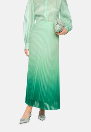 Frau trägt einen grünen Ombre-farbenen, plissierten Midirock, eine transparente Langarmbluse, hält eine silberne Clutch und trägt verzierte spitze Pumps.