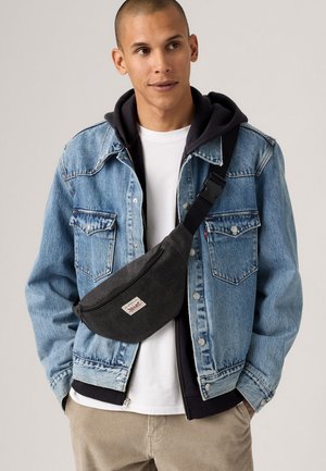 Man wearing blue denim jacket over black hoodie and white shirt, with black crossbody bag and beige pants, looking to the side.