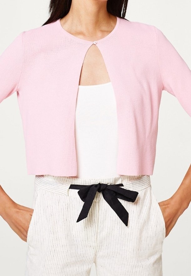 Cardigan corto de color rosa claro con escote redondo, que presenta un recorte en forma de ojo de cerradura y un botón decorativo. Se lleva sobre una camiseta blanca y unos pantalones a rayas.