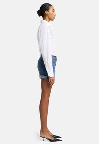 Chemise blanche à manches longues avec boutons, shorts en denim et mules noires à talons pointus. La tenue présente un design ajusté et des bords frangés.