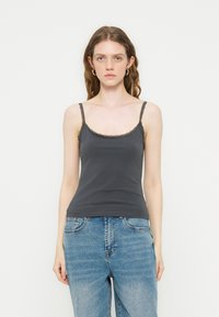 Νέα γυναίκα με ελεύθερα κυματιστά μαλλιά φοράει σκουρόγκρι κορμάκι με δαντέλα και ανοιχτό μπλε ψηλόμεσο τζιν, στέκεται μπροστά σε λευκό φόντο.