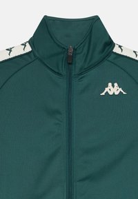 Tirkizna zip jakna s visokim ovratnikom, glatke teksture i bijelim detaljima s logom na prsima. Detaljno šivanje uz rubove.
