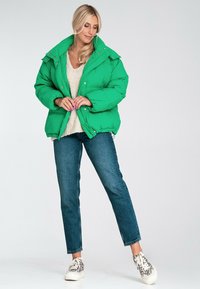 Zielona kurtka puchowa z wysokim kołnierzem i zatrzaskami, w połączeniu z kremowym, grubym swetrem, ciemnoniebieskimi dżinsami i wzorzystymi sneakersami.