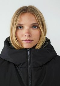 Czarna kurtka puchowa z wysokim kołnierzem, zamkiem błyskawicznym z przodu i gładką teksturą. Model ma proste blond włosy i neutralny wyraz twarzy.