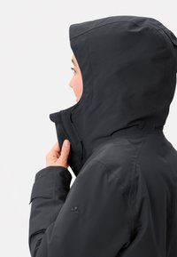 Donkergrijs waterdicht jack met kap. Kenmerken zijn onder andere verstelbare kraag, ritsafsluiting en logoaccent op de mouw. Zachte textuur.